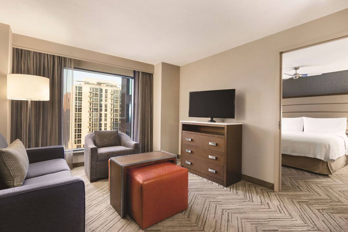 Une chambre d’hôtel équipée d’un canapé, d’un fauteuil, d’un pouf, d’une télévision, d’une commode et d’un lit.
