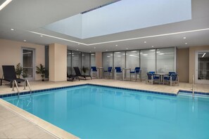 A modern indoor pool with clear glass walls, blue chairs, and a skylight.