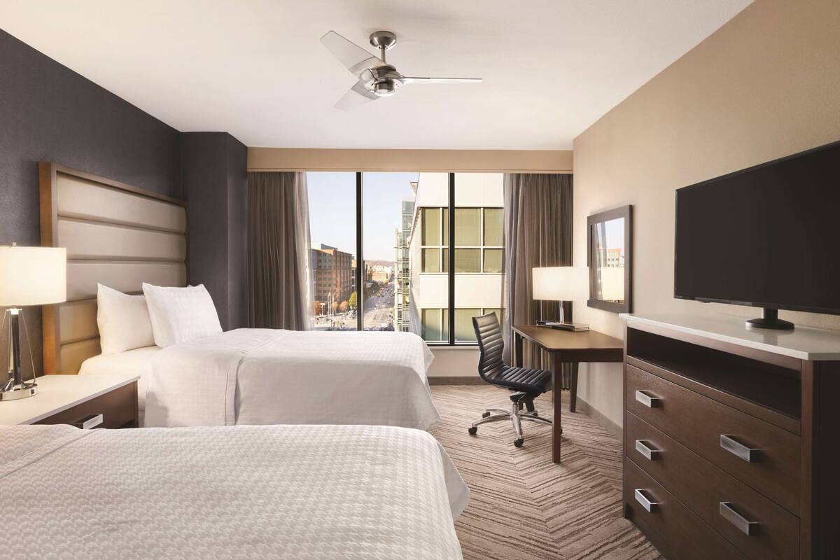 Habitación de hotel con dos camas, un escritorio, una silla, un televisor y un amplio ventanal con vistas a la ciudad.
