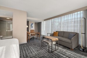 Habitación de hotel con cama, sofá, escritorio y un amplio ventanal con cortinas.