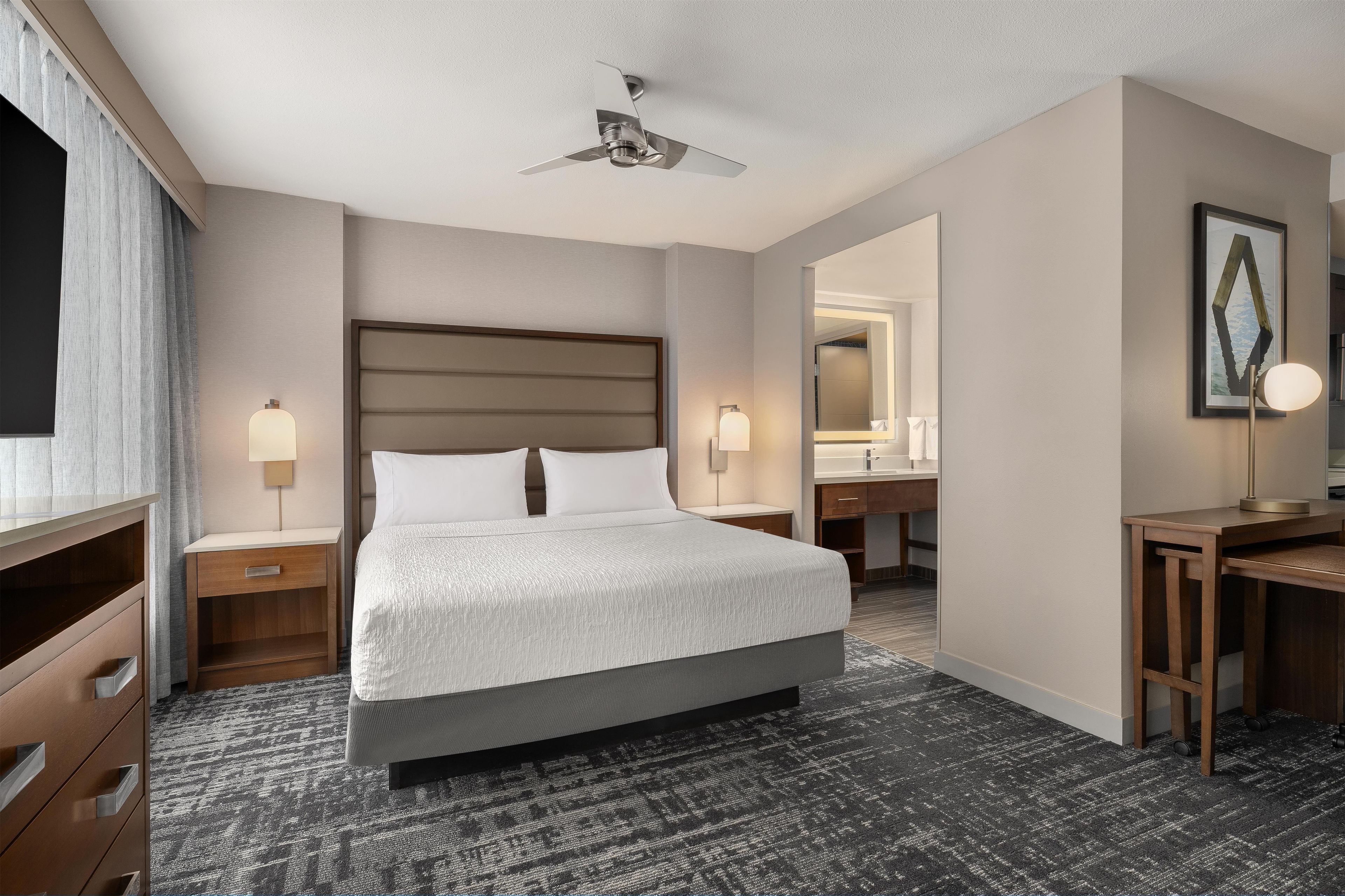 Une chambre d’hôtel avec un grand lit, des tables de chevet, un bureau, une chaise, un ventilateur de plafond et un grand miroir.