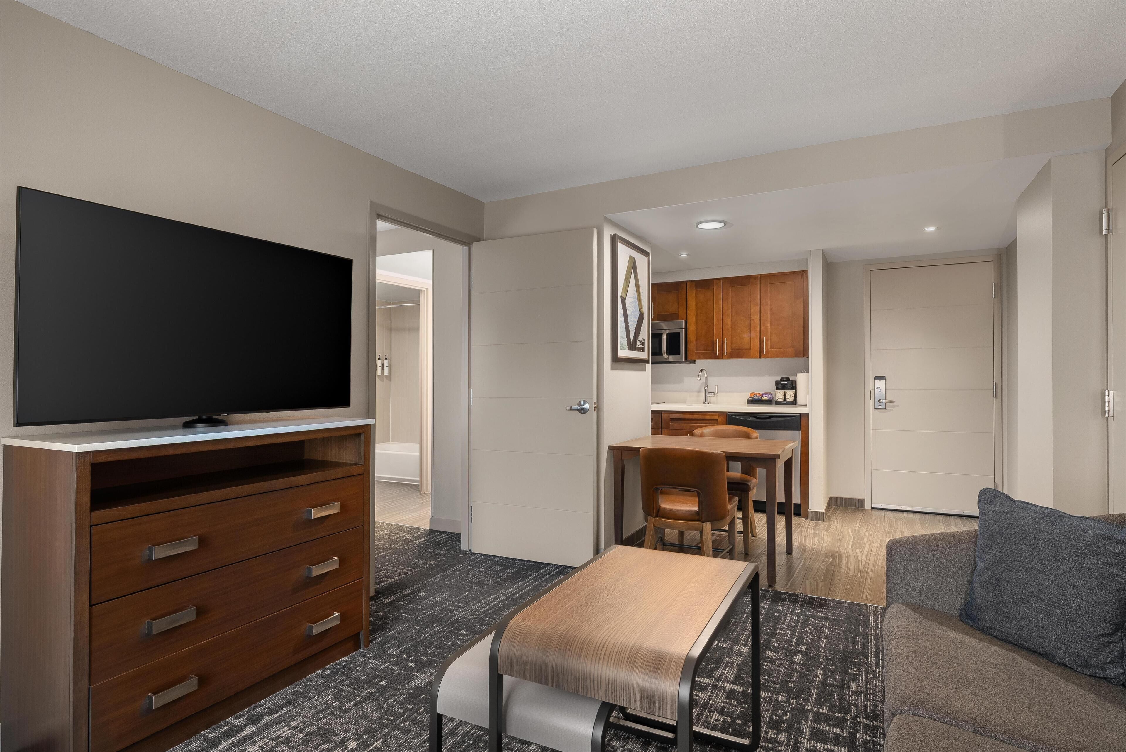 A modern hotel room with a flat-screen TV, wooden furniture, a dining area, and a kitchenette.
