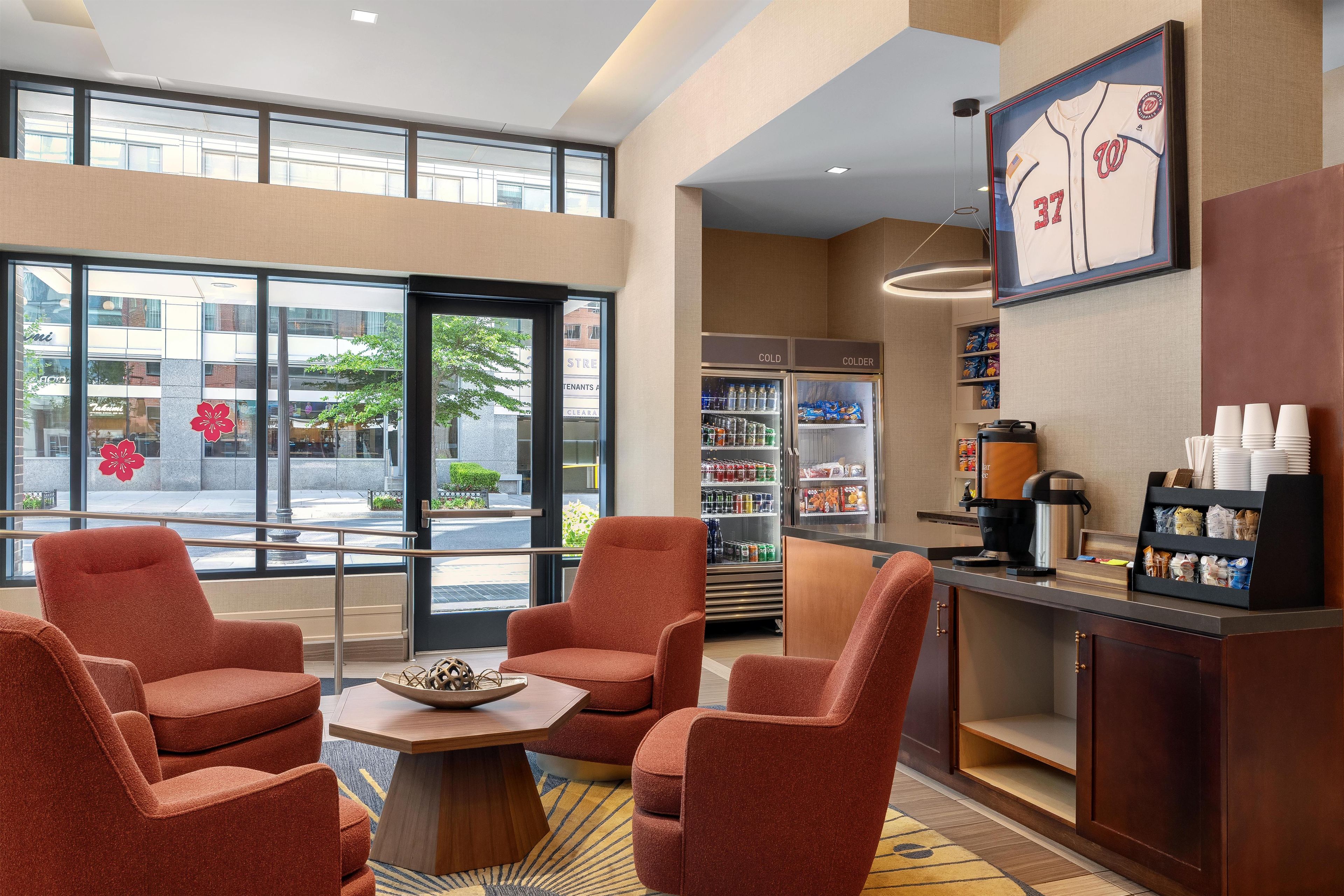 A hotel lobby with seating, a coffee station, a refrigerator, and a large window with a view of the street.