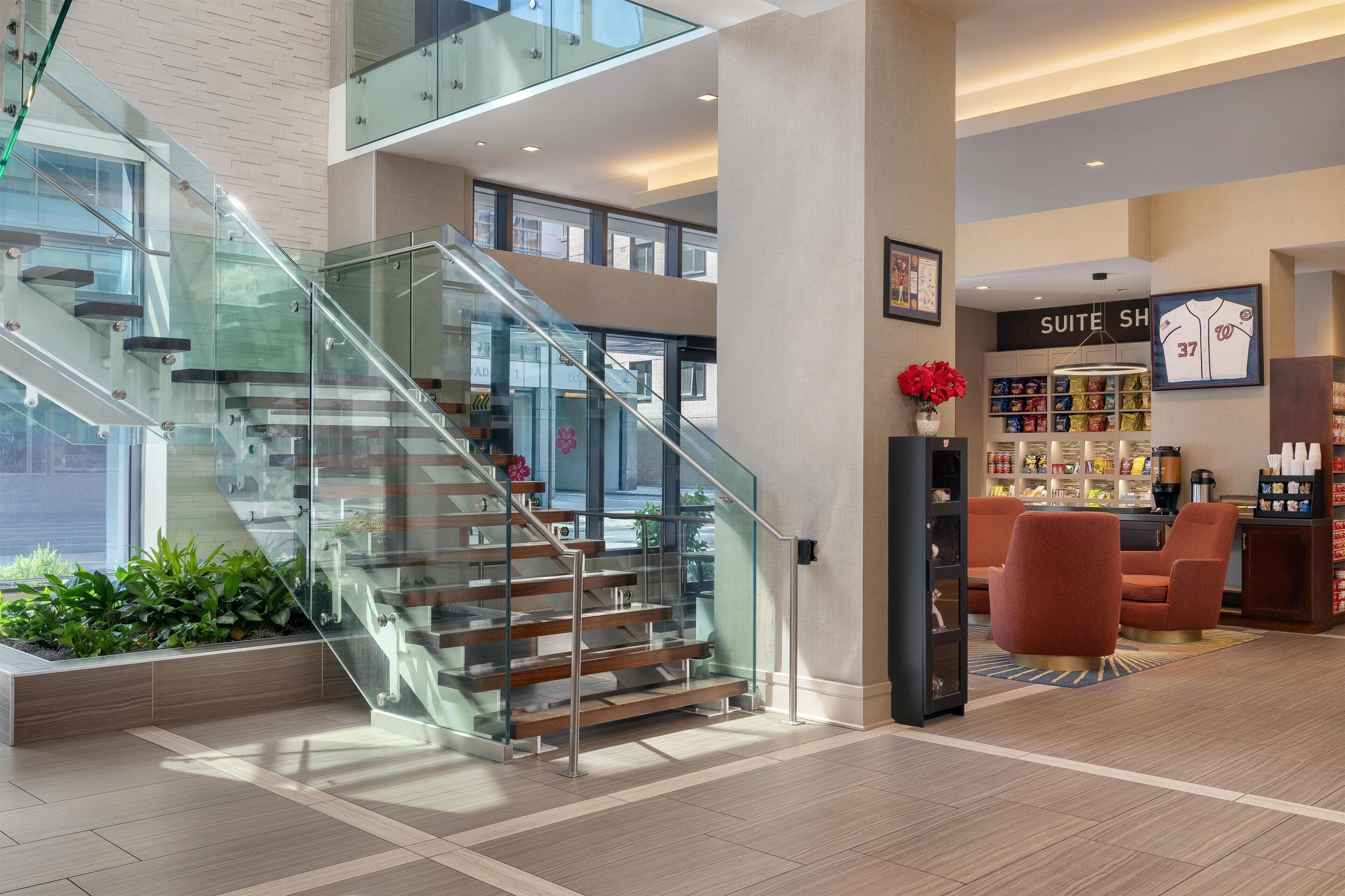 A modern hotel lobby with a glass staircase, a visible 'SUITE SH' sign, and a seating area with red chairs.