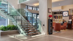 Un moderno vestíbulo de hotel con una escalera de cristal, un letrero visible que dice 'SUITE SH' y una zona de estar con sillas rojas.
