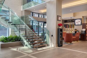 Un moderno vestíbulo de hotel con una escalera de cristal, un letrero visible que dice 'SUITE SH' y una zona de estar con sillas rojas.