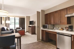 Habitación de hotel con kitchenette, zona de comedor con mesa y sillas, sofá, televisor y un ventanal con cortinas.