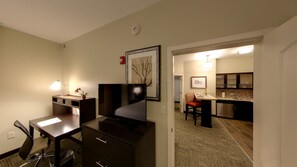 Quarto de hotel com escrivaninha, cadeira, TV e cozinha compacta.