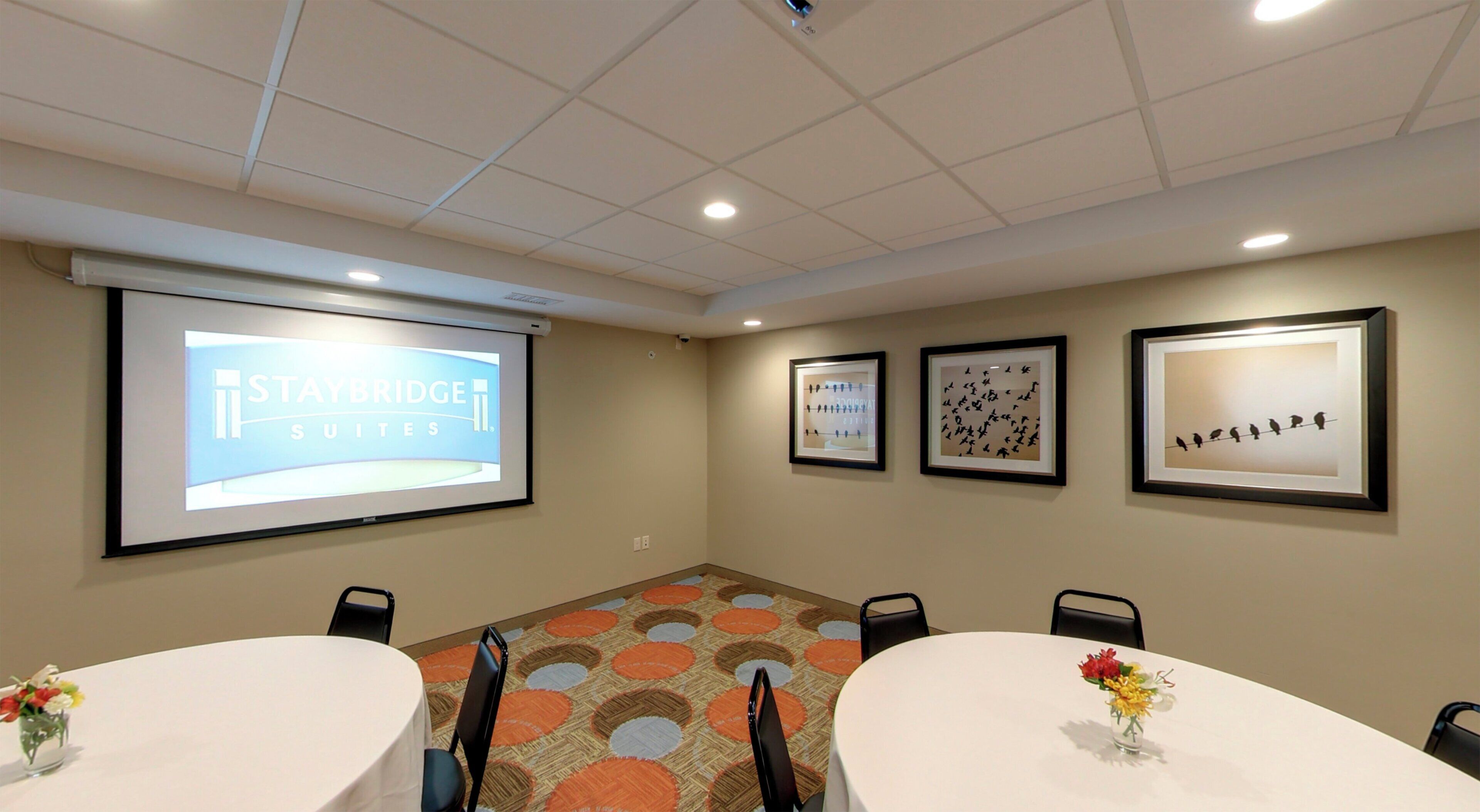 Une salle de conférence équipée d’un écran de projection sur lequel est affiché le nom « STAYBRIDGE SUITES », de deux œuvres d’art encadrées et d’une moquette à motifs circulaires.