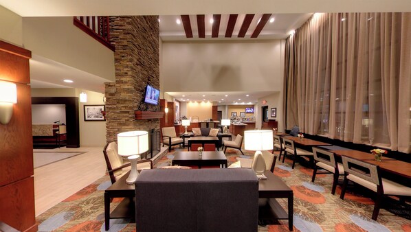 Um saguão de hotel com parede de destaque em pedra, uma televisão montada acima de uma lareira e uma área de jantar com mesas e cadeiras.