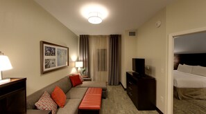 Quarto de hotel com sofá, uma mesinha com abajur, televisão em um móvel, cama e um quadro na parede.