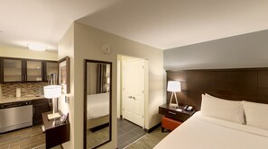 Quarto de hotel com cama, mesa de cabeceira, luminária e vista para o banheiro e cozinha.