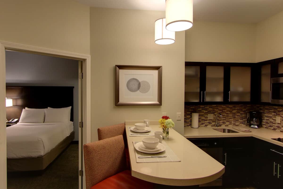 Une chambre d’hôtel comprenant un lit, un petit coin salon avec une table préparée pour deux personnes, une kitchenette et une œuvre d’art encadrée au mur.