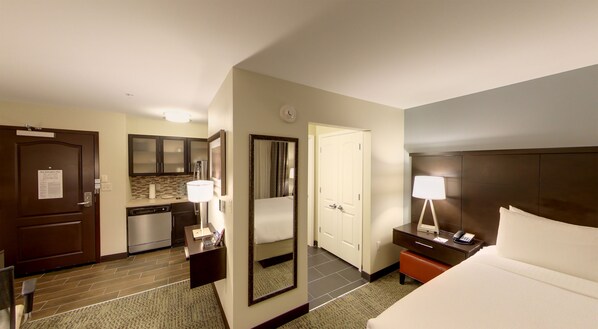 Quarto de hotel com cama, mesa de cabeceira, luminária, armário, espelho e área de cozinha compacta.