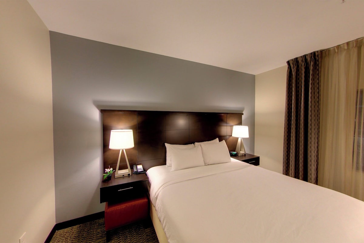 Quarto de hotel com uma cama grande, duas mesas de cabeceira com abajures, uma cabeceira escura e uma janela com cortinas.
