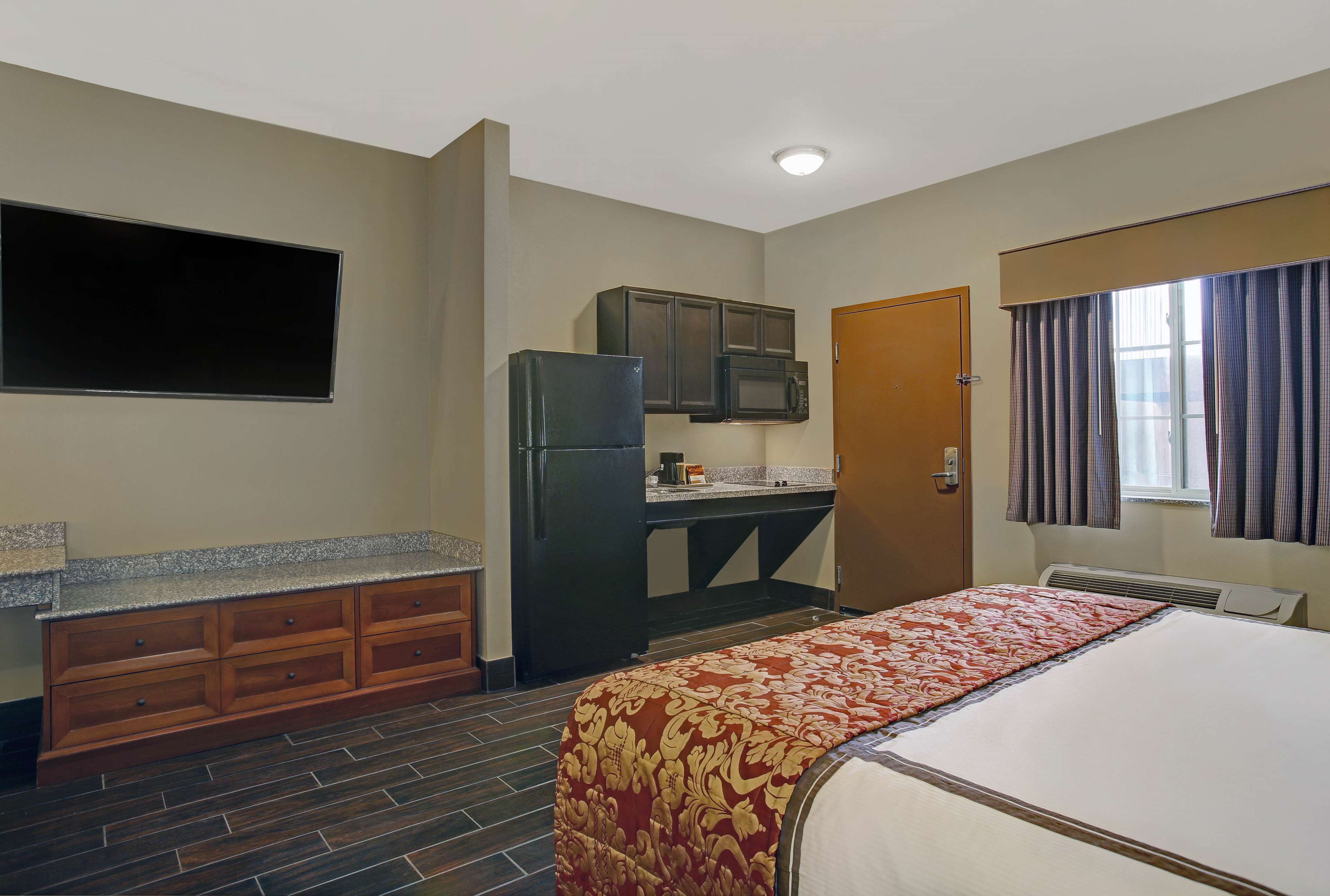 A compact hotel room with a kitchenette, a bed with a patterned bedspread, and a window with curtains.