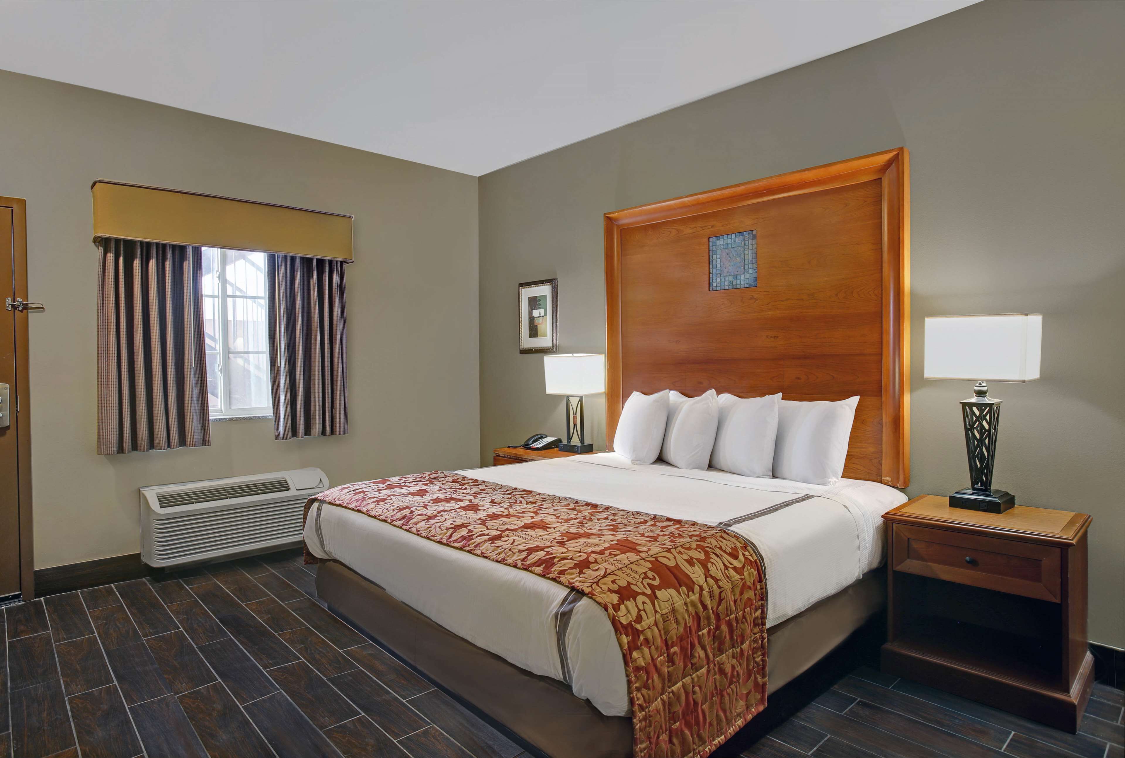 A hotel room with a large bed, a wooden headboard, a bedside table with a lamp, a nightstand, a window with curtains, a wall-mounted heater, and a framed picture on the wall.