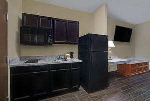 Cocina compacta con muebles oscuros, un microondas, un refrigerador, un lavamanos y un televisor de pantalla plana montado en la pared.