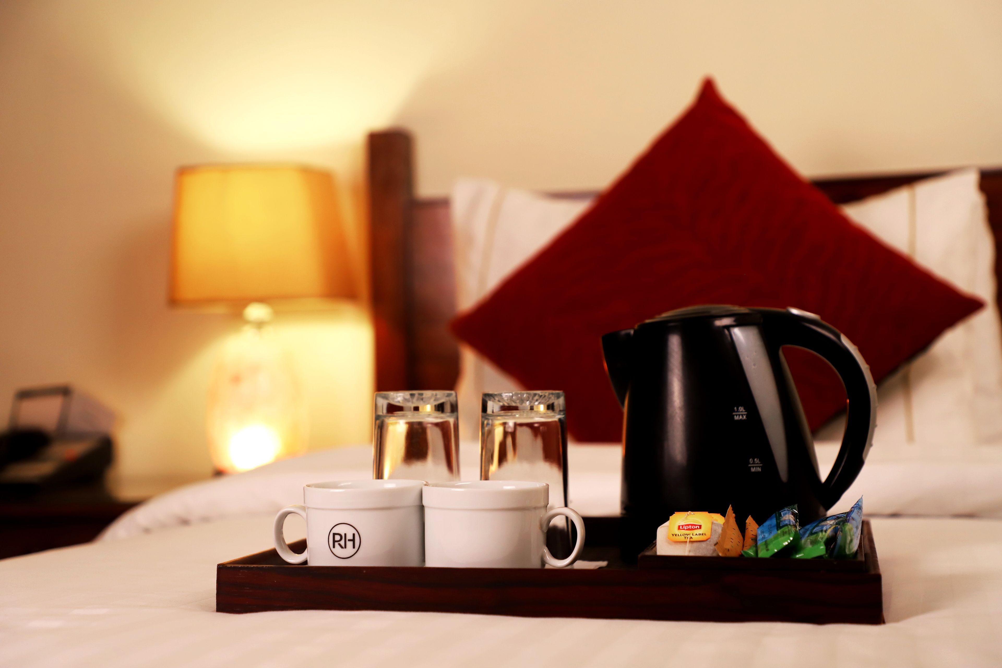 Habitación de hotel con cama, una bandeja con dos tazas blancas, una cafetera negra y una almohada roja.