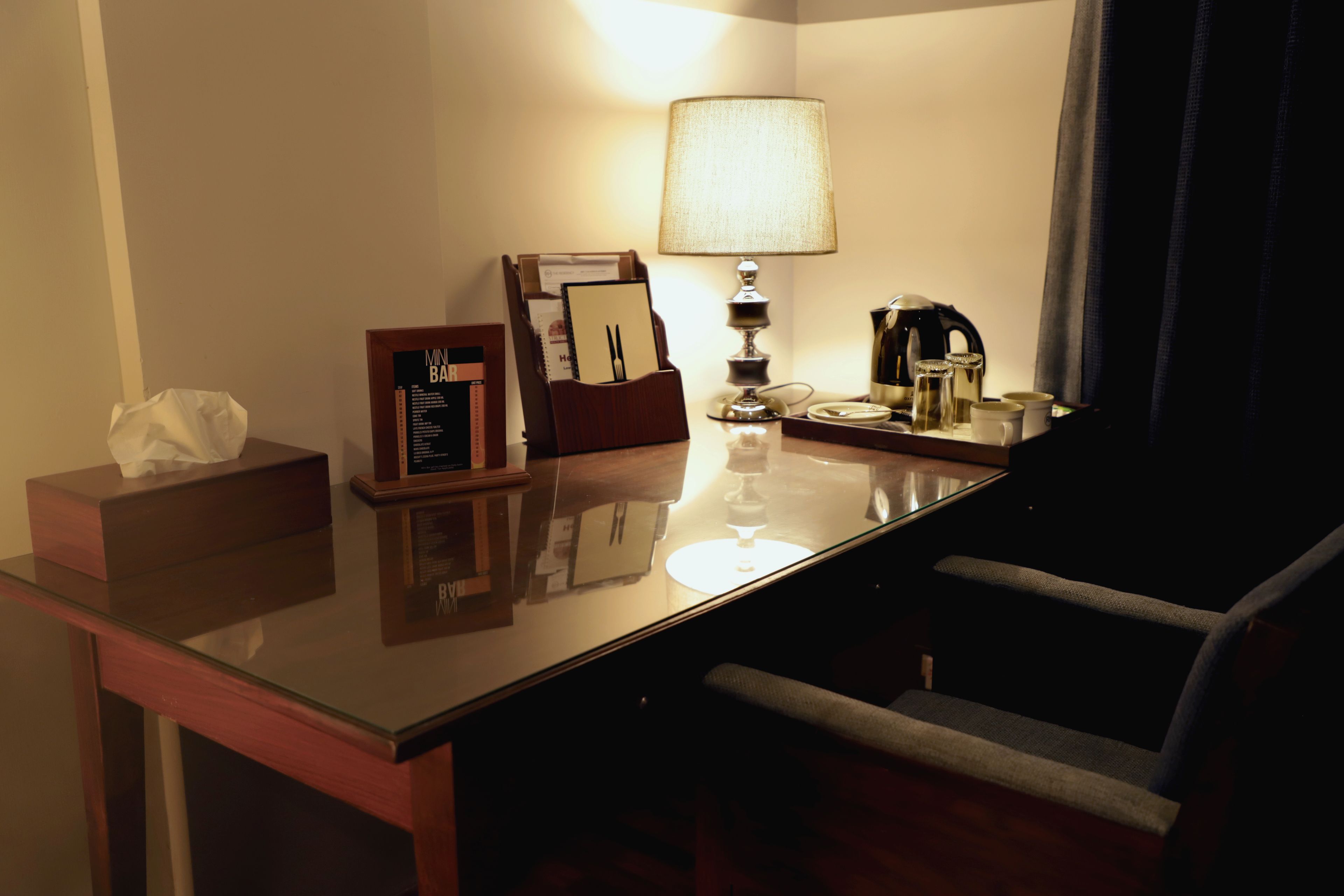 A hotel room with a glass desk, a lamp, a coffee maker, a tissue box, and a framed menu.