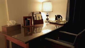 A hotel room with a glass desk, a lamp, a coffee maker, a tissue box, and a framed menu.