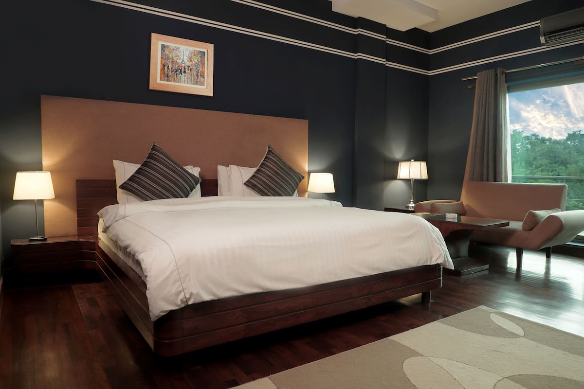 A spacious bedroom with a large bed, two bedside tables with lamps, a chair, and a window with a view of trees.