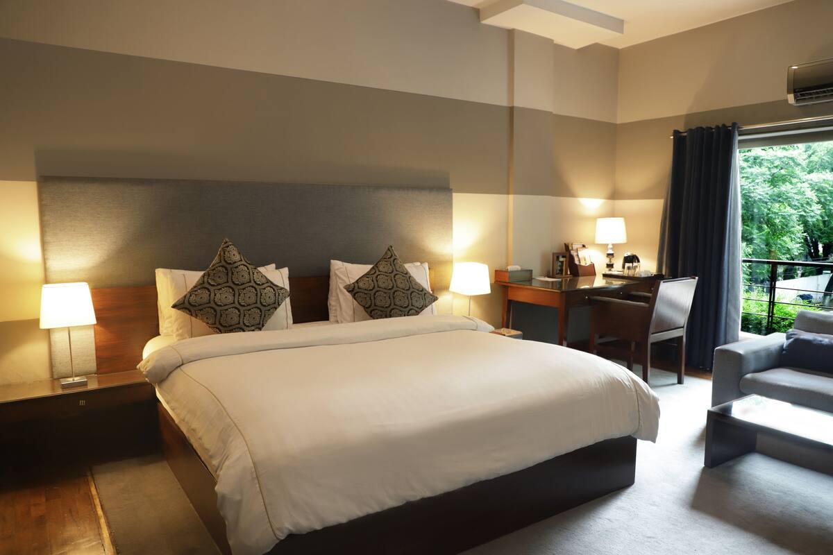 A hotel room with a large bed, two bedside tables with lamps, a desk with a chair, and a balcony with a view of greenery.
