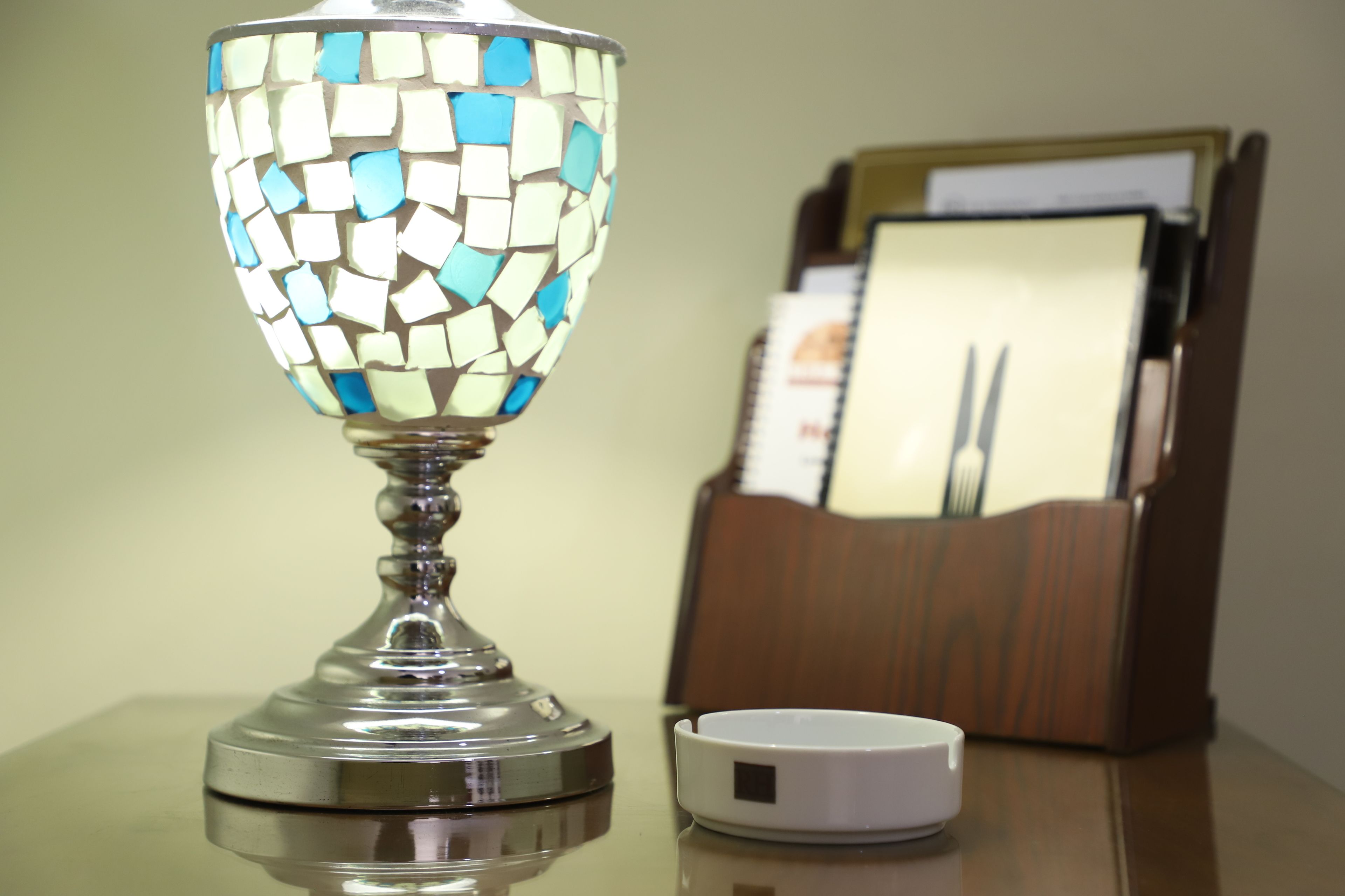 Una lámpara de vidrio con diseño tipo mosaico sobre una mesa, junto a un cenicero blanco y una mesita de madera con un portalápices.