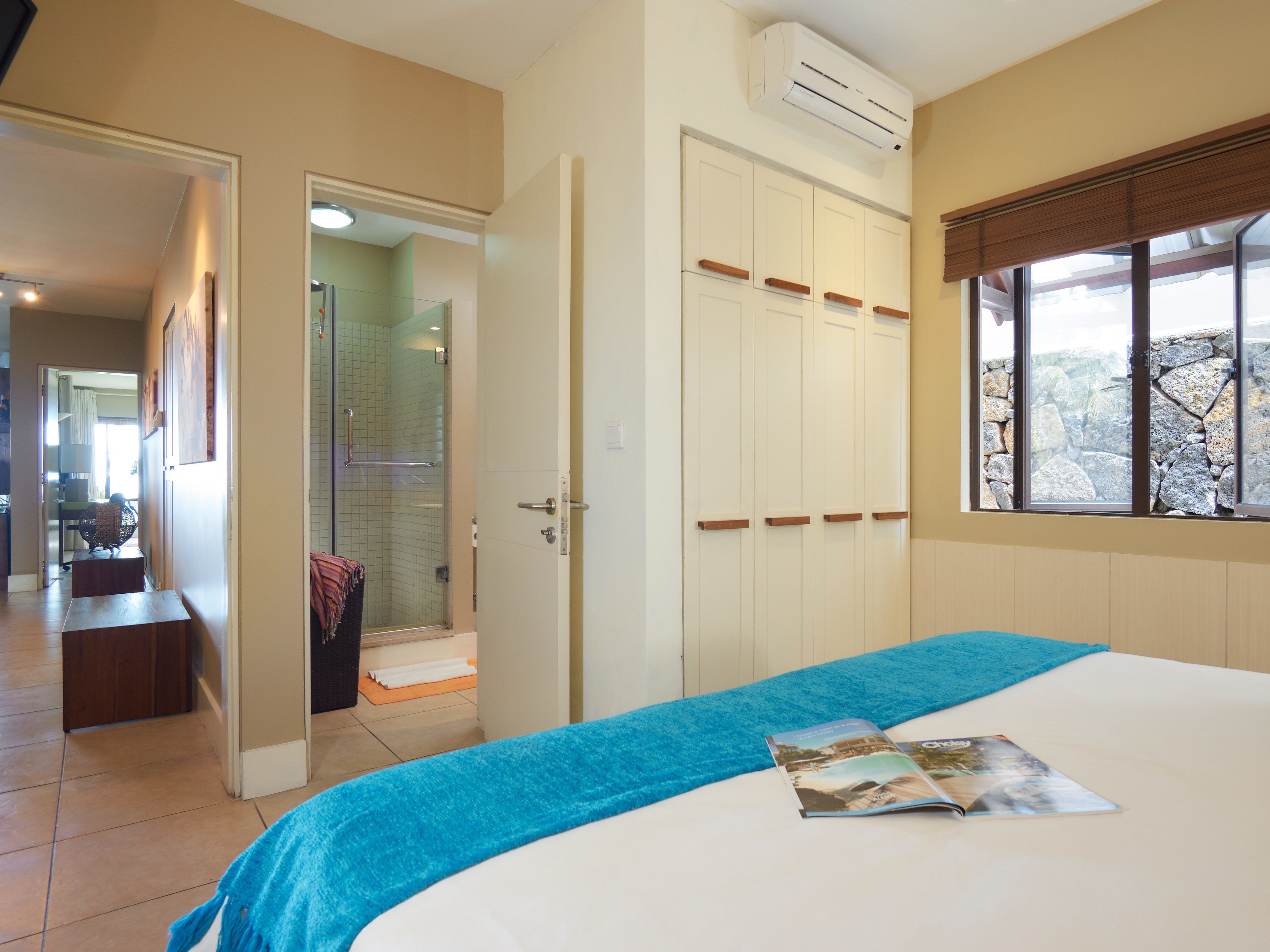 A hotel room with a bed, a window with a stone wall view, a bathroom with a glass door, and a hallway leading to another room.
