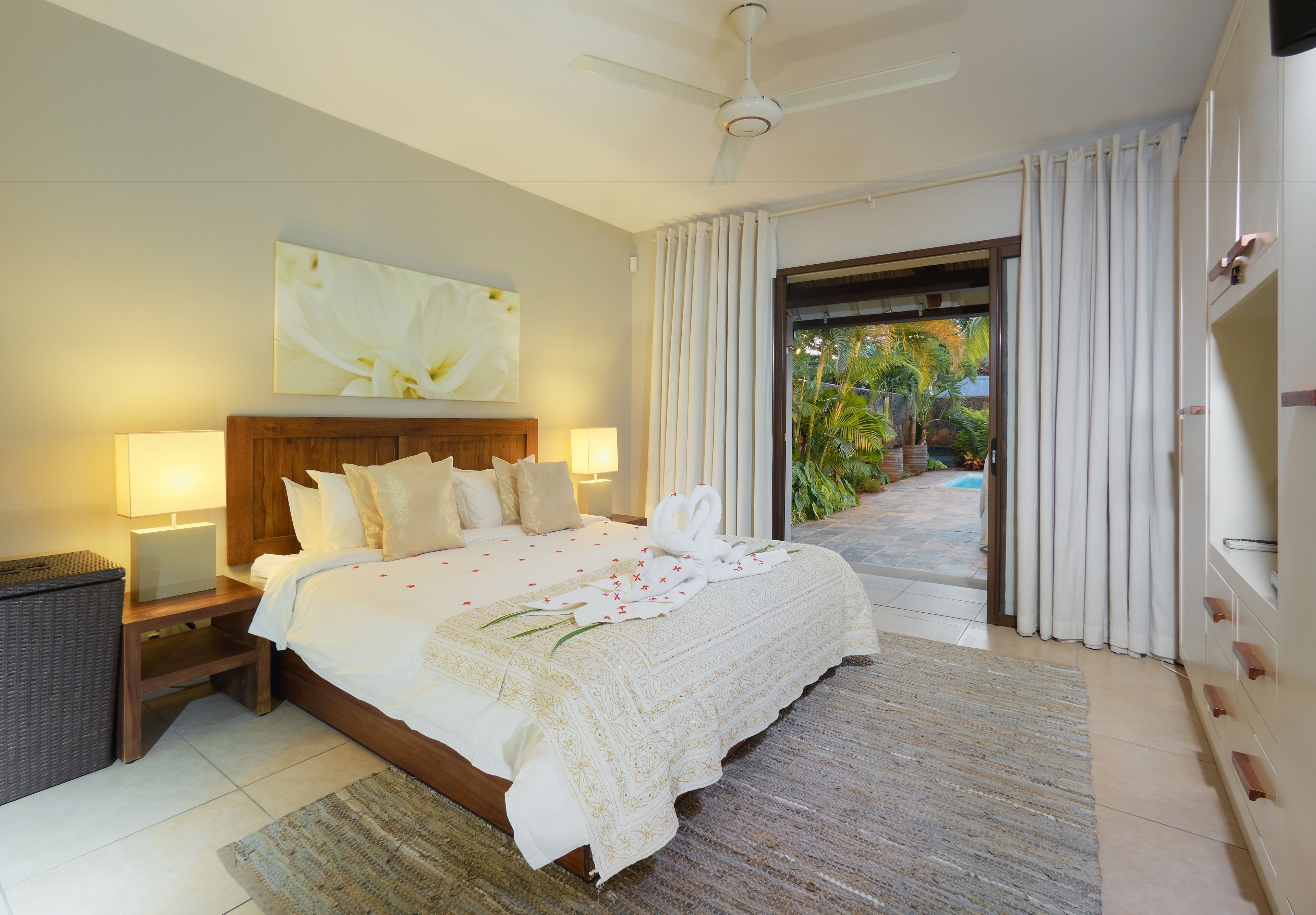 Ein geräumiges Schlafzimmer mit einem großen Bett, zwei Nachttischlampen, einem Deckenventilator und Blick auf einen Garten durch eine offene Tür.