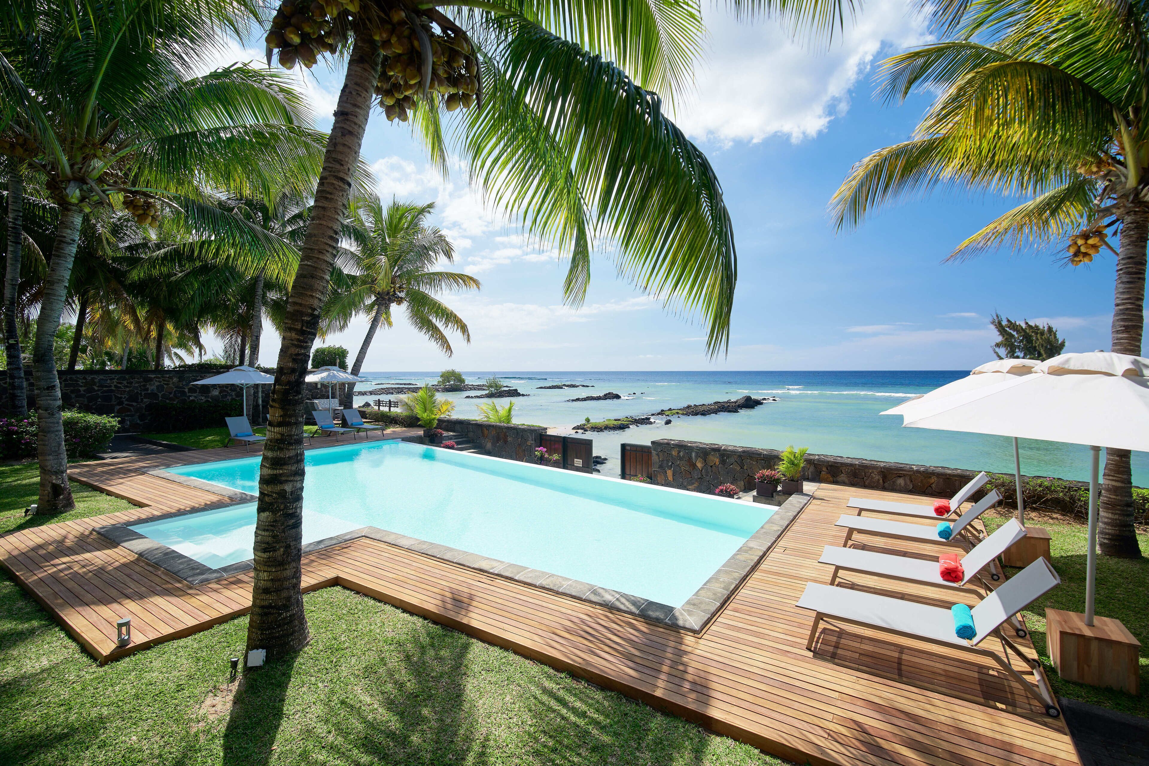 Un espace piscine agrémenté d’une terrasse en bois, de chaises longues et de palmiers.