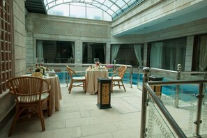 A spacious indoor area with a pool, featuring a high ceiling, large windows, and a seating area with tables and chairs.