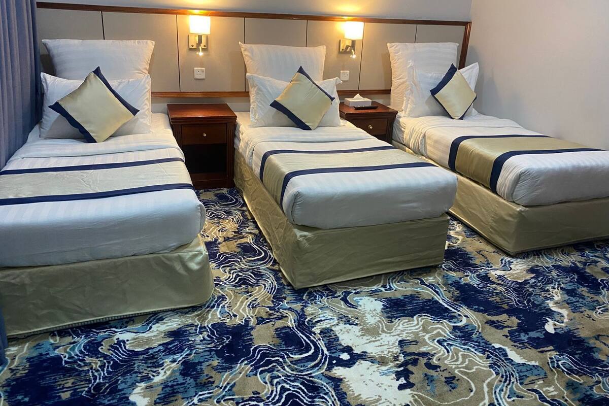 A hotel room with three single beds, each with a headboard, bedside tables, and lamps. The room has a patterned carpet and a plain wall with a light switch above the beds.