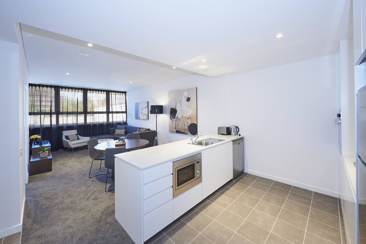 A modern kitchen with a central island, built-in oven, and microwave. There is a dining area with a round table and chairs, and a living area with a sofa and coffee table.