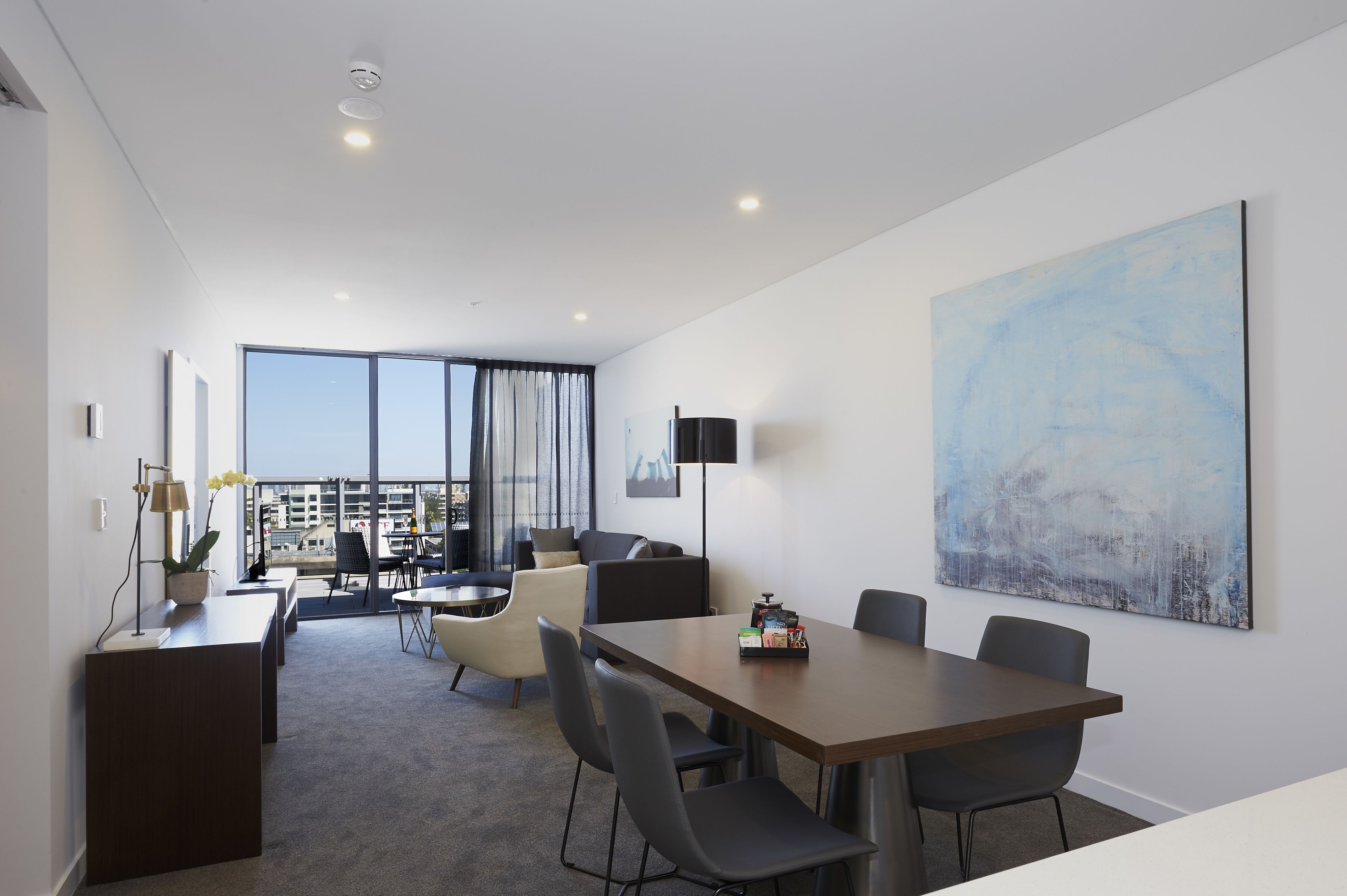 A modern hotel room with a dining area, a large window with curtains, a flat-screen TV, and a large abstract painting on the wall.