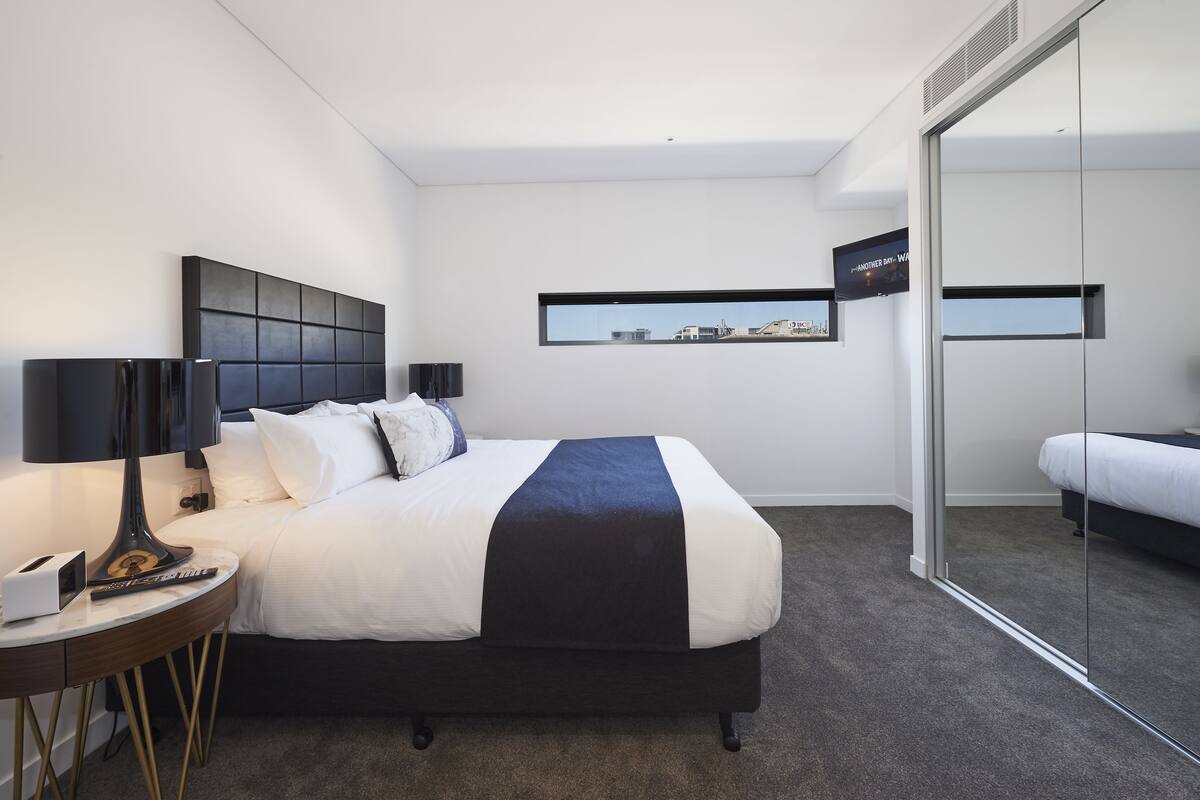 A modern hotel room with a large bed, a bedside table with a lamp, a flat-screen TV, and a city view through a window.