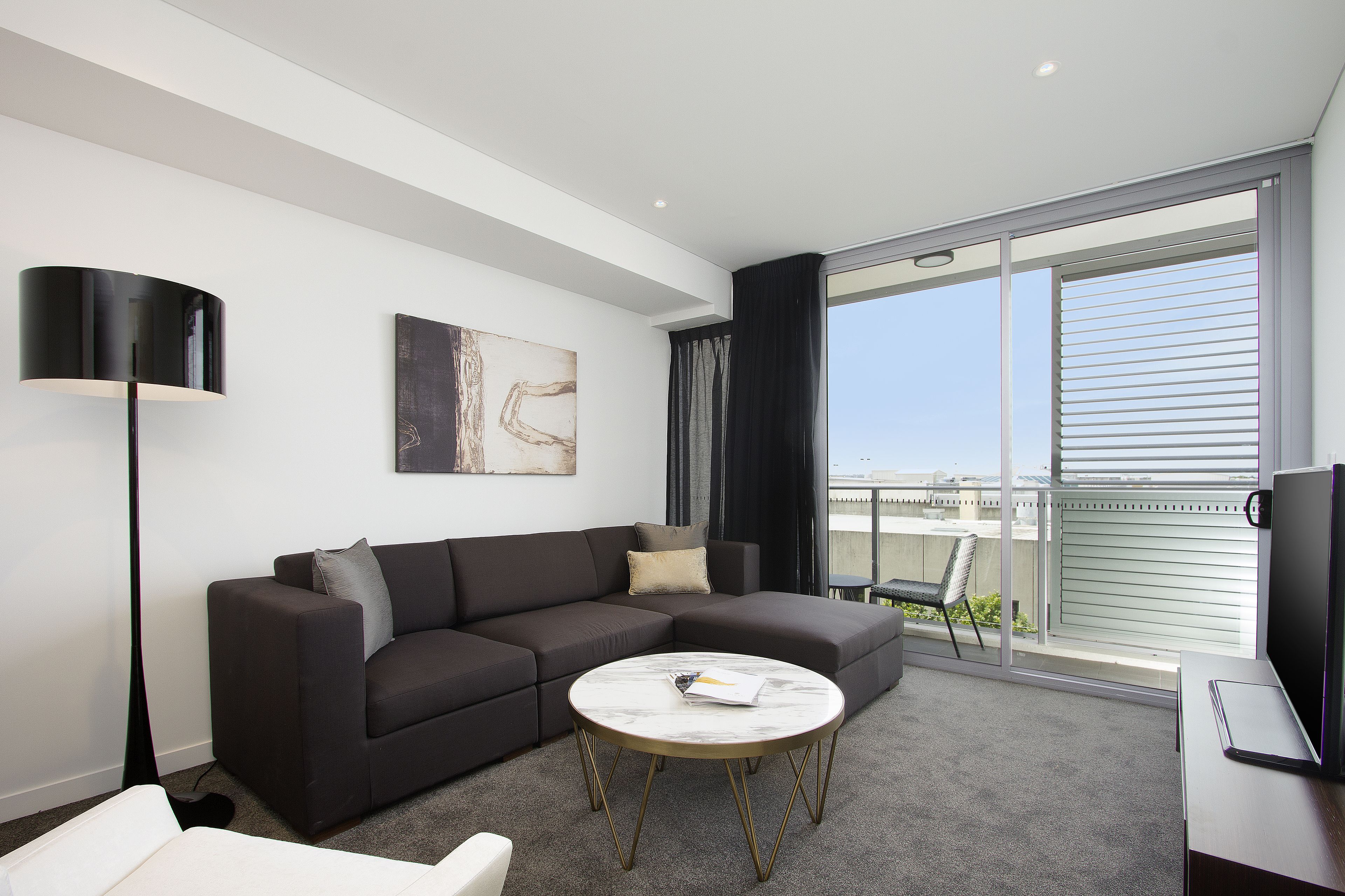 A modern living room with a dark grey sofa, a round coffee table, and a floor lamp. There is a large window with blinds, and a painting on the wall.