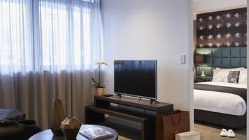 A modern hotel room with a large flat-screen TV, a bed with a tufted headboard, a small round table with a vase and magazine, and a window with sheer curtains.