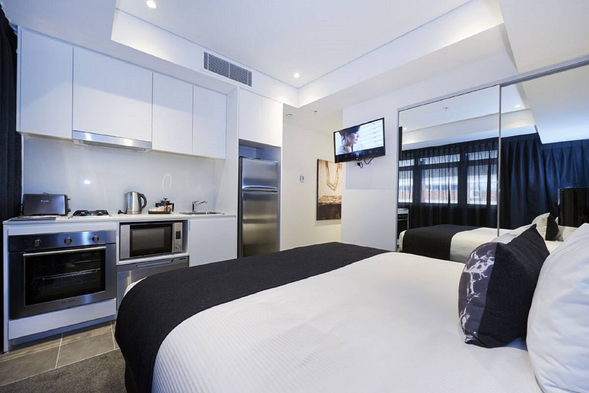 A modern hotel room with a kitchenette, a bed with black and white bedding, a flat-screen TV mounted on the wall, and a large window with curtains.