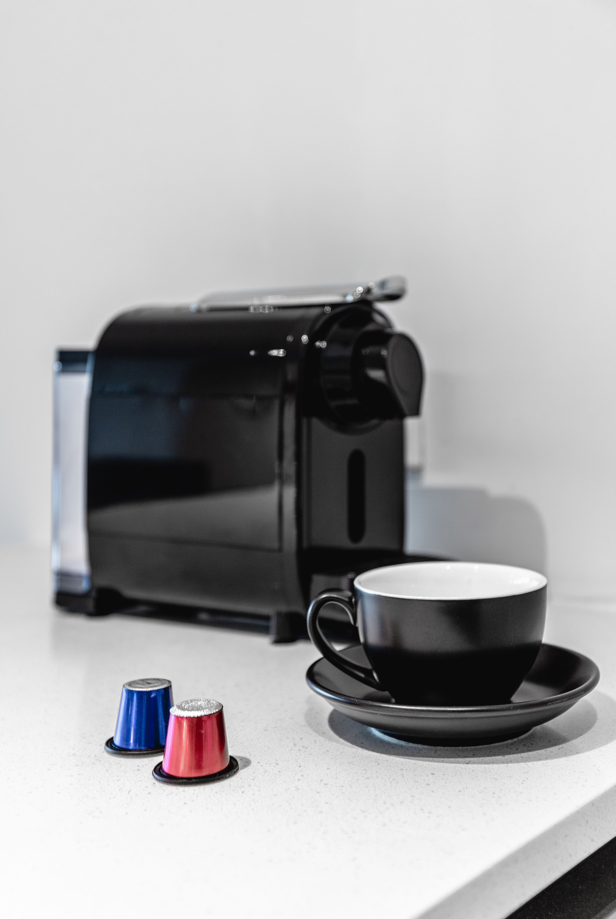 A coffee maker, a black cup with a saucer, and two capsules on a countertop.