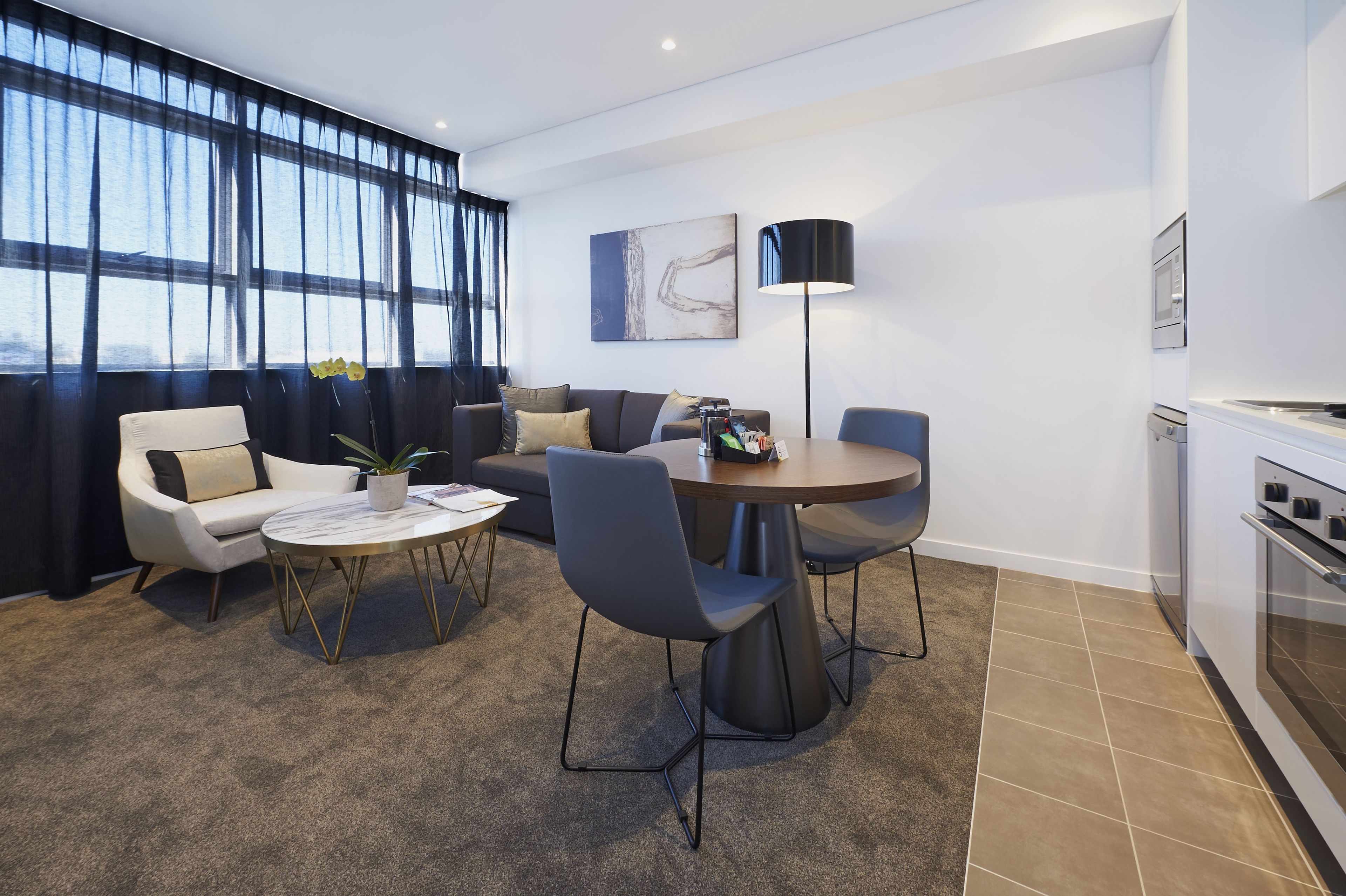 Una sala de estar moderna con sofá, sillones, una mesa de centro y una mesa de comedor. Hay un ventanal con cortinas, un cuadro en la pared y una zona de cocina con horno.