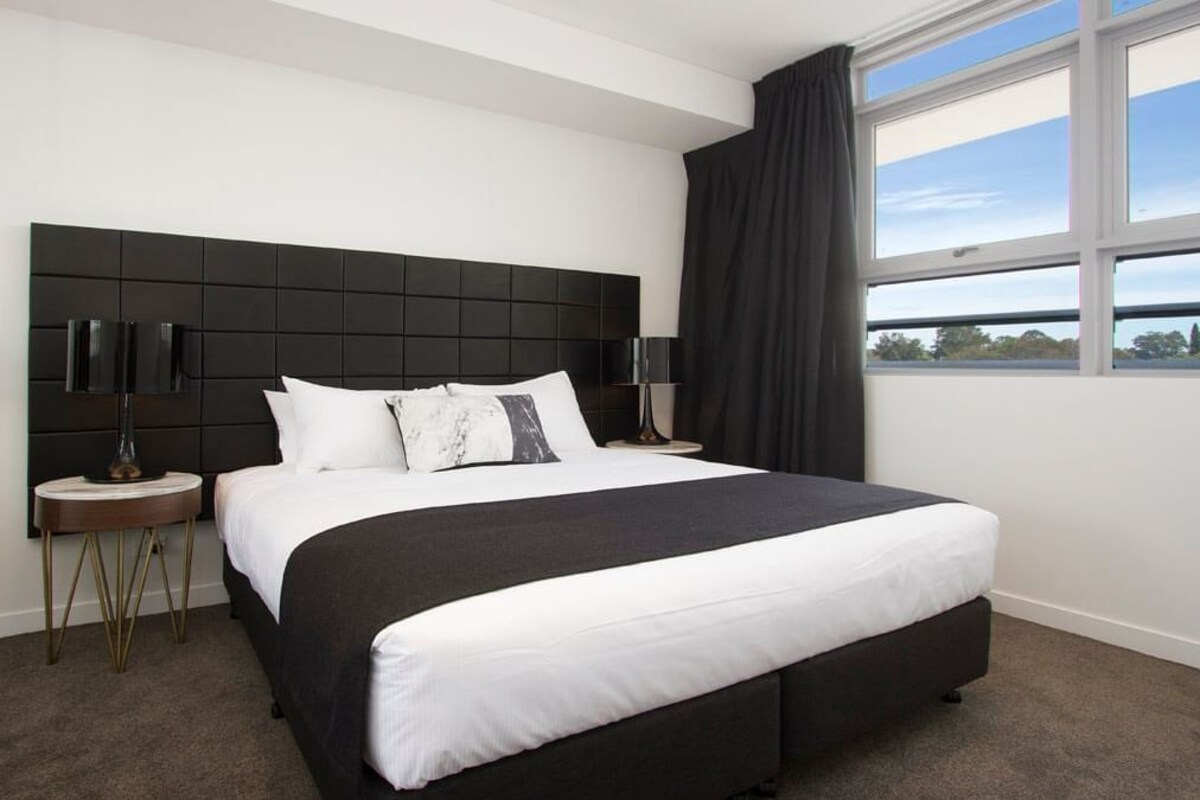 Habitación de hotel moderna con una cama grande, dos mesitas de noche, un cabecero negro y vistas al exterior a través de una ventana.