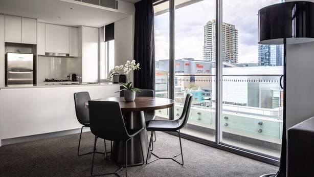 Una cocina moderna con gabinetes blancos, un refrigerador de acero inoxidable y una zona de comedor con una mesa redonda y sillas negras. Hay una planta en maceta sobre la mesa, y la habitación tiene ventanas amplias que ofrecen vistas de la ciudad.