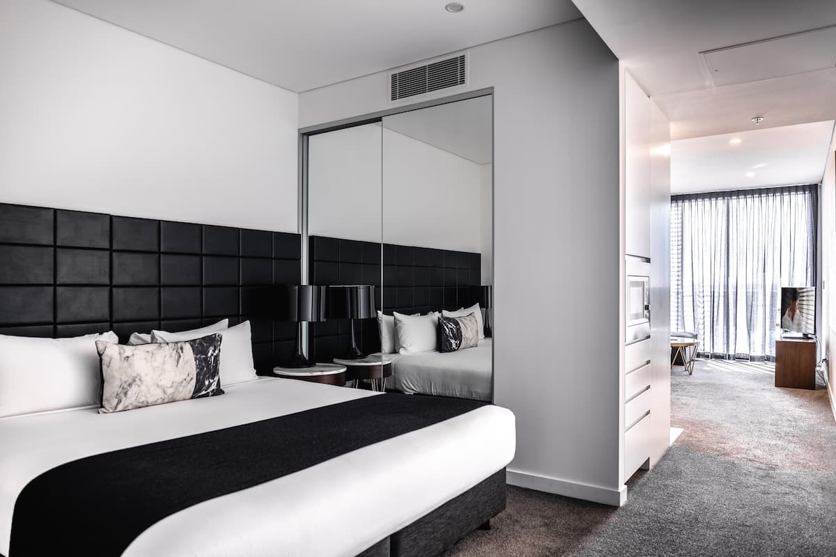 A modern hotel room with a large bed, a black headboard, a bedside table with a lamp, and a large mirror.