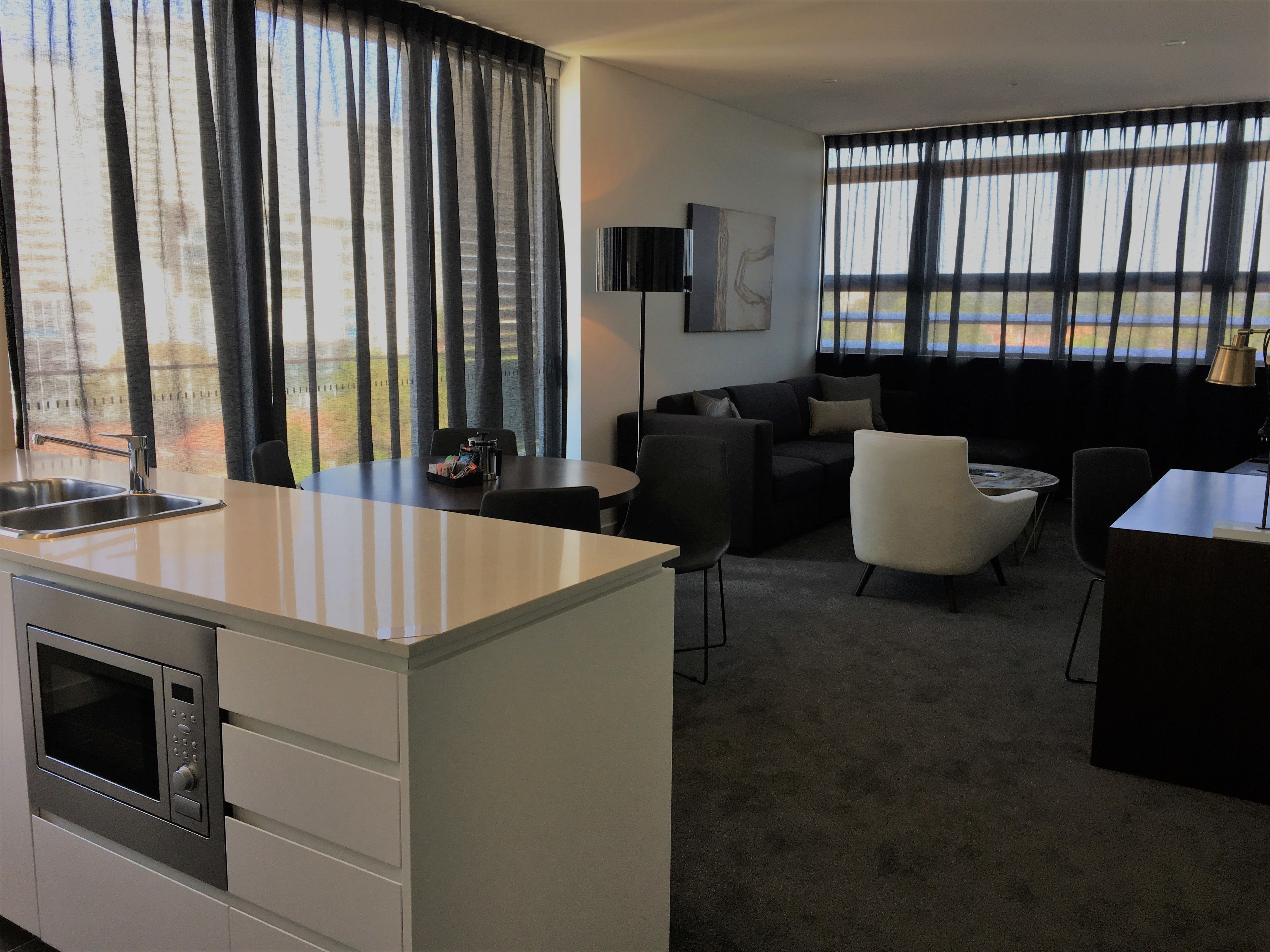 A modern hotel room with a kitchenette, a sofa, a dining table, and large windows with curtains.