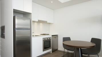 A modern kitchen with white cabinetry, a stainless steel refrigerator, a built-in oven, and a dishwasher. There is a round table with two chairs and a gas stove.
