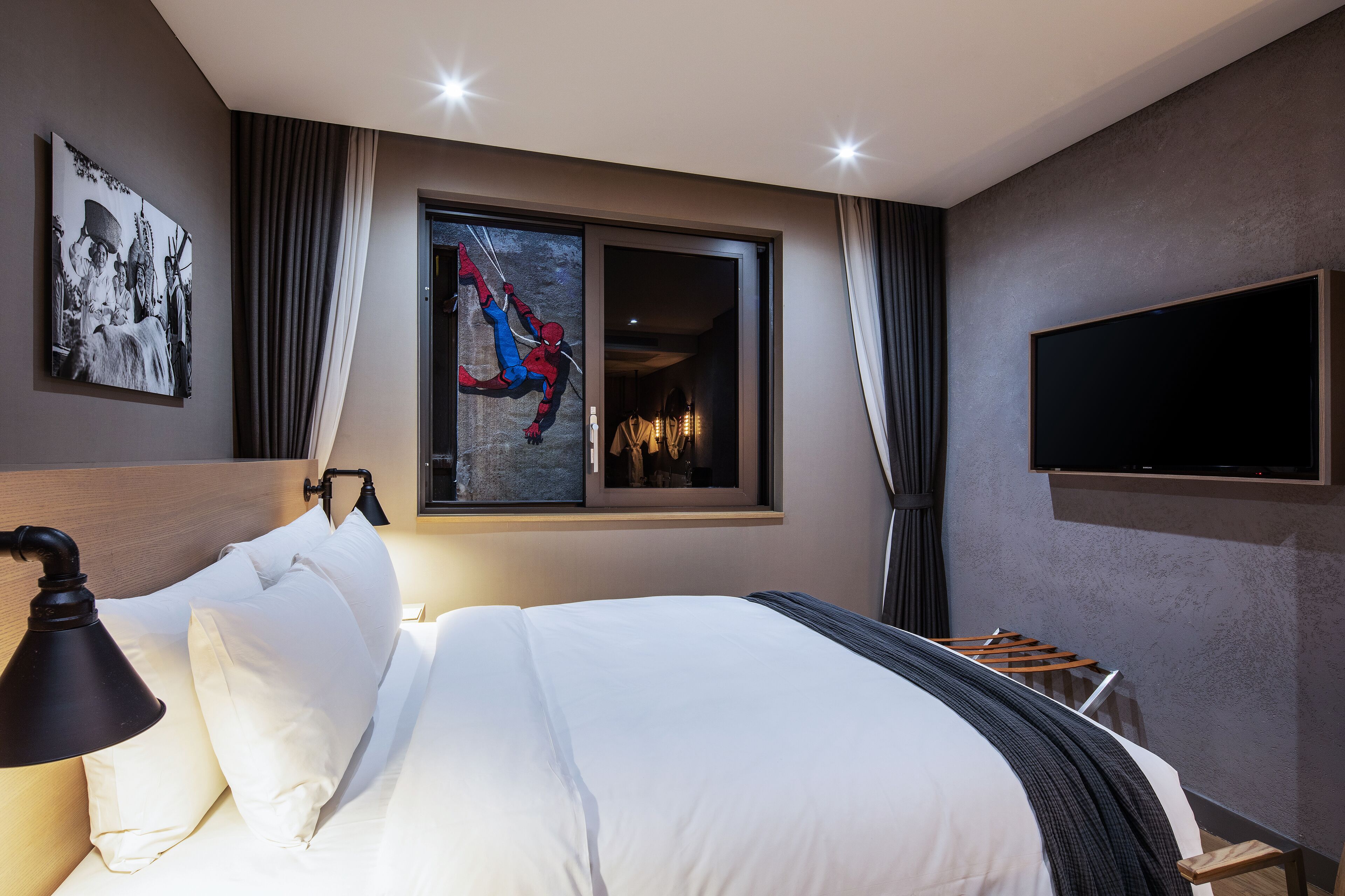A modern hotel room with a large bed, a wall-mounted TV, a window with a view, and a painting on the wall.