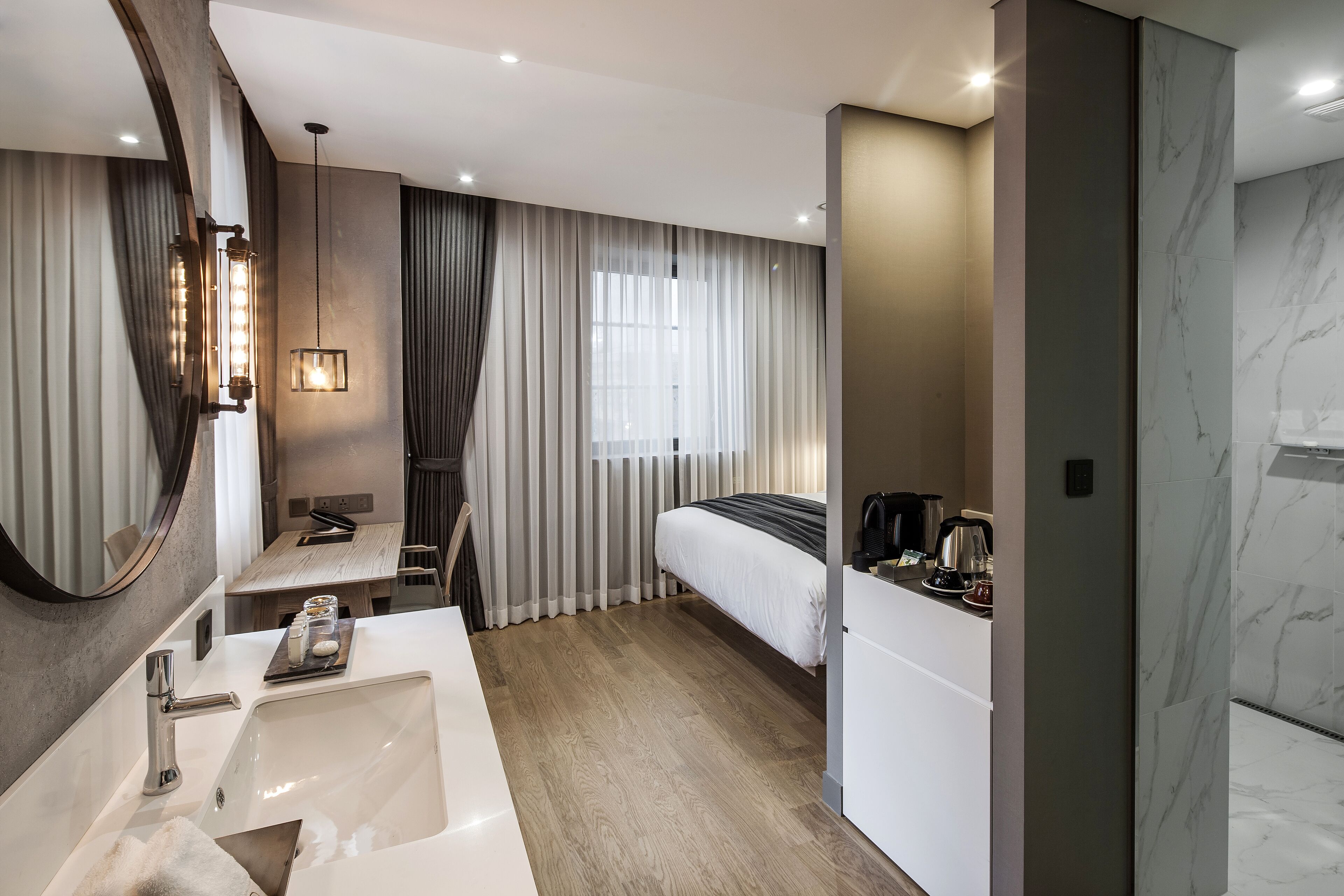 A modern hotel room with a large bed, a separate desk with a chair, a bathroom with a large mirror, and a sink.