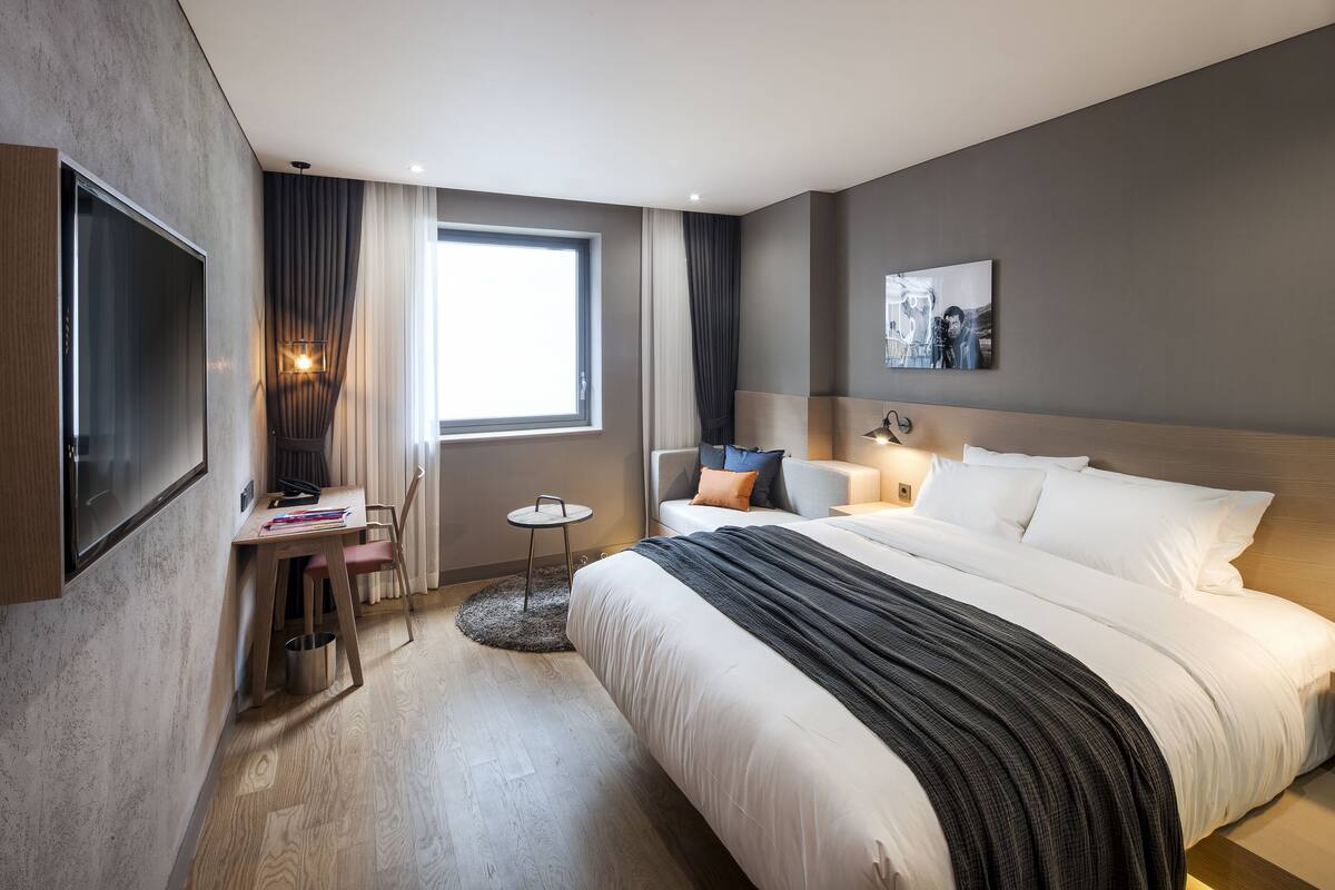 A modern hotel room with a large bed, a desk with a chair, a small round table, and a TV mounted on the wall.