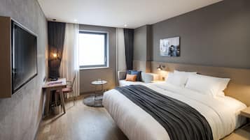 A modern hotel room with a large bed, a desk with a chair, a small round table, and a TV mounted on the wall.